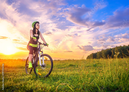 Young sporty woman at sunset on a bicycle. Sporty girl on bike stopped to enjoy the moment at beautiful sunset. Lens flare against the sky background © marmoset
