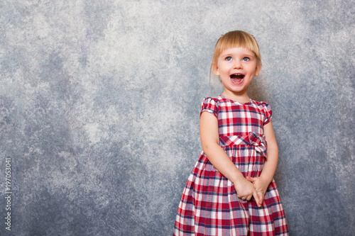 A tiny sweet girl with blond hair wearing a red checkered dress. She makes faces on a gray background. Wide angle and copy space. Child day. © AnastazjaSoroka