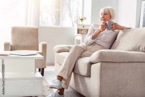 Relaxing in morning. Beautiful elderly woman sitting on the couch in her living room, drinking coffee and contemplating something