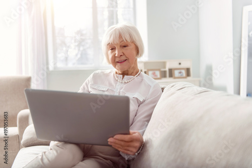 Experienced businesswoman. Pleasant upbeat elderly woman sitting on the sofa and reading a business email on her laptop while smiling © zinkevych