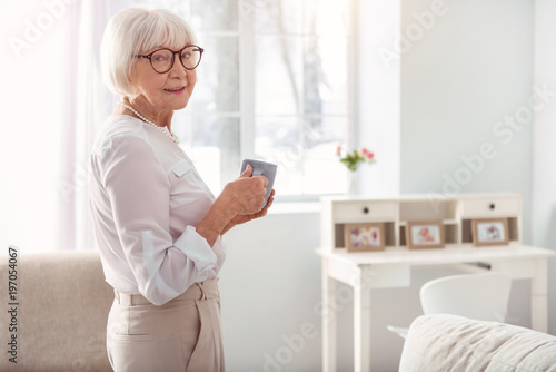 Caffeine lover. Pleasant pretty elderly woman standing half-turned in the living room and posing for the camera while holding a cup of coffee © zinkevych