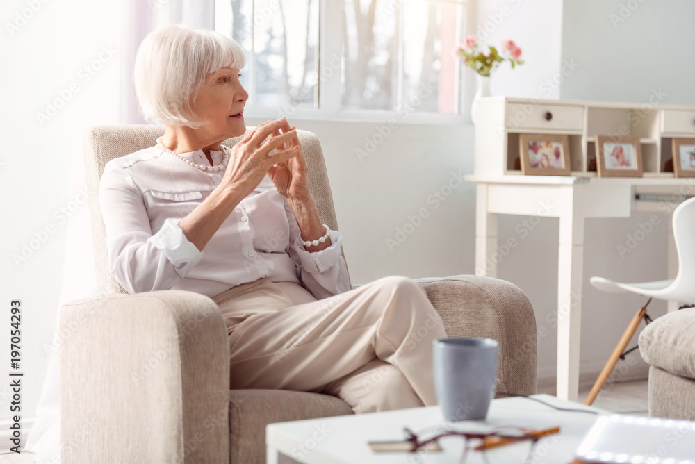 Pensive mood. Pretty elderly lady sitting in the armchair and thinking about something while pressing the fingertips on both hands together Pensive mood. Pretty elderly lady sitting in the armchair and thinking about something while pressing the fingertips on both hands together