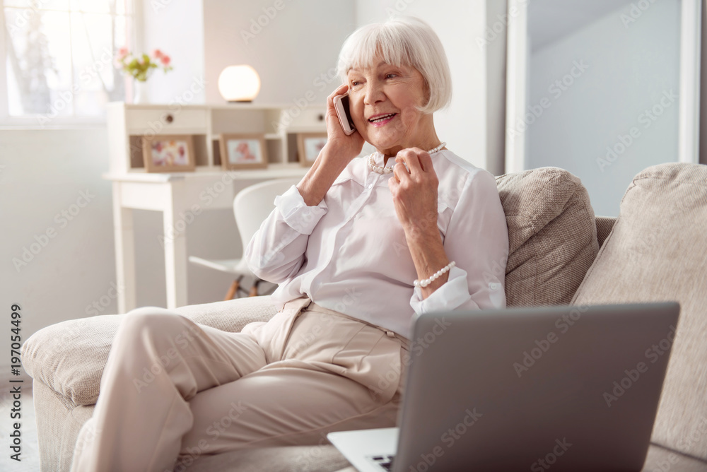 Pleasant conversation. Cheerful elderly lady sitting on the sofa in the living room and talking on the phone while smiling happily Pleasant conversation. Cheerful elderly lady sitting on the sofa in the living room and talking on the phone while smiling happily