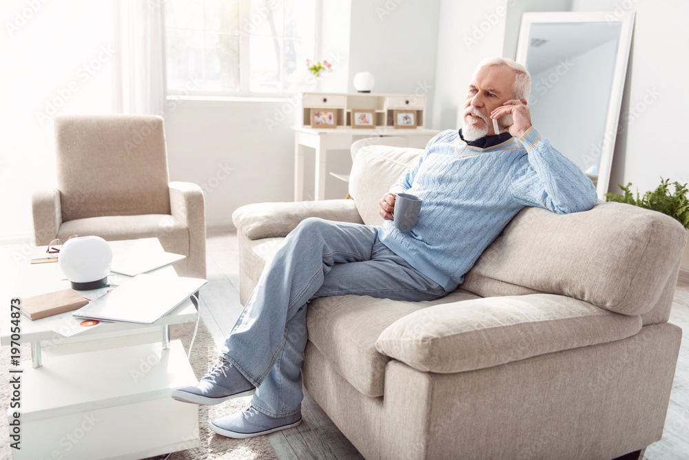 Important discussion. Pleasant senior man sitting on the couch in the living room and having a serious conversation on the phone while drinking coffee
