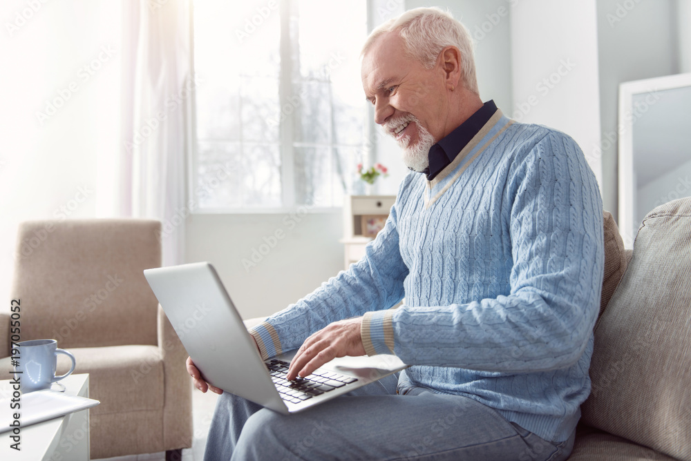 Mood-boosting chat. Upbeat senior man sitting on the couch and typing an email on the laptop while smiling happily Mood-boosting chat. Upbeat senior man sitting on the couch and typing an email on the laptop while smiling happily