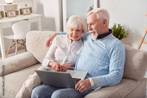 Convenient purchase. Happy elderly couple sitting on the couch in the living room and choosing a new laptop in the online store together