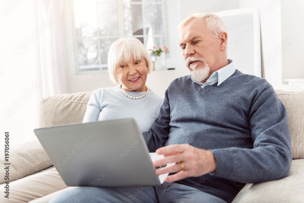Keeping themselves updated. Pleasant elderly husband and wife sitting on the couch and scrolling the newsfeed on the laptop, being slightly dissatisfied with the news