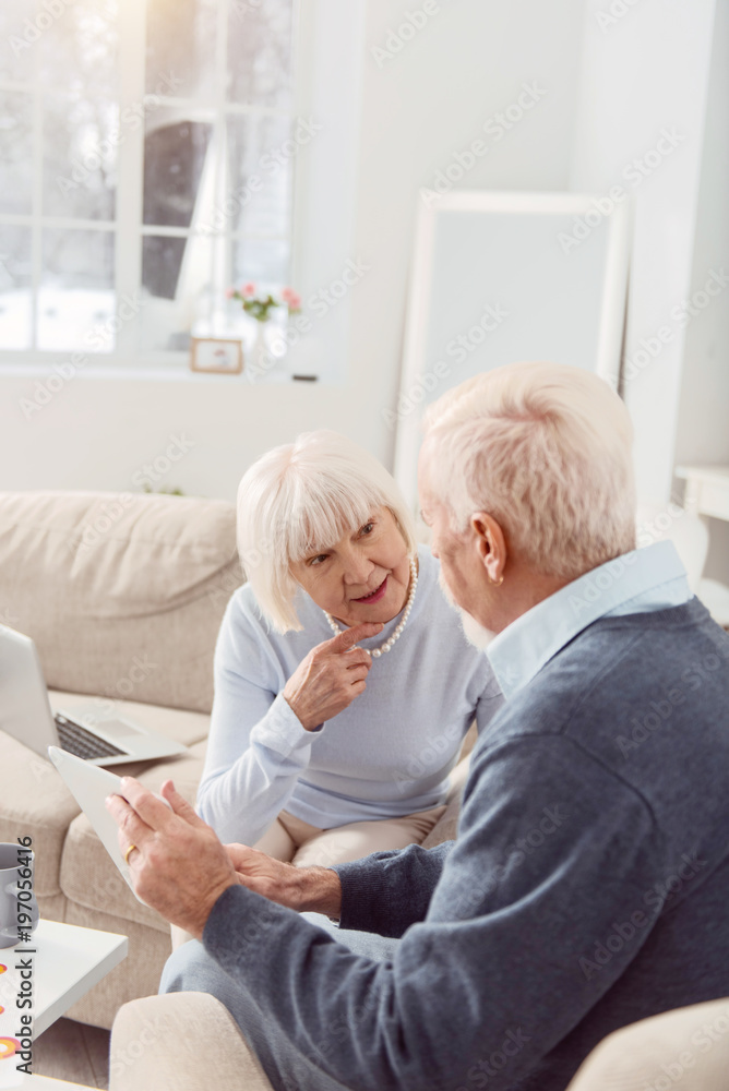 Obraz premium Discussion of intellectuals. Pleasant senior couple sitting in the living room, reading the news online on tablet and talking about world events