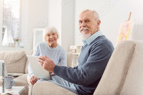 Mood-boosting message. Joyful elderly man and his beloved wife sitting in the living room and watching a video message from their children on tablet