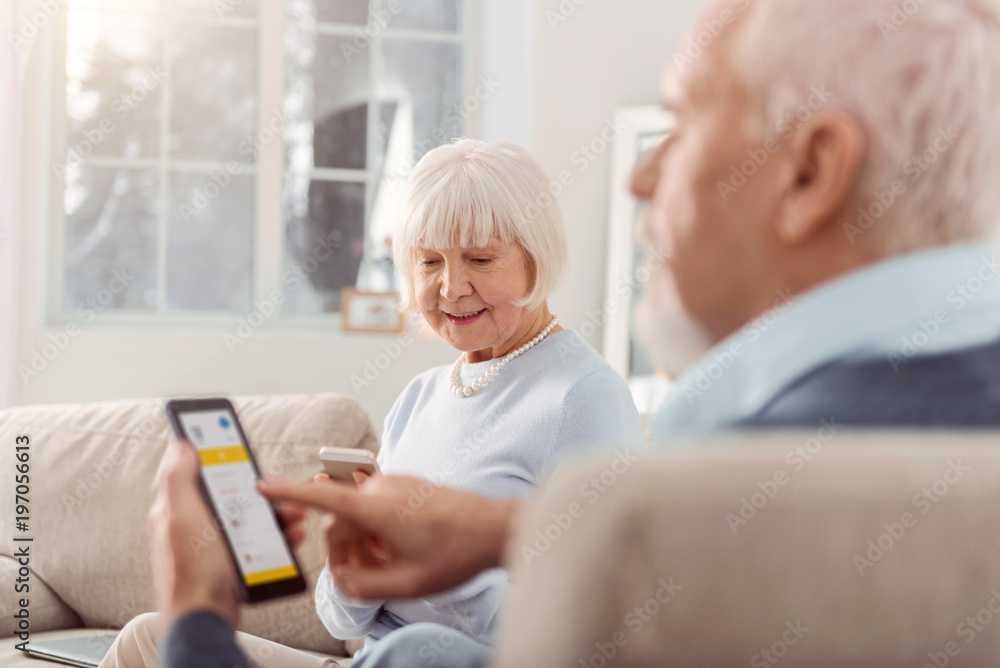 Useful technologies. Pleasant elderly couple sitting in the living room and being focused on their mobile phones while using the applications Useful technologies. Pleasant elderly couple sitting in the living room and being focused on their mobile phones while using the applications