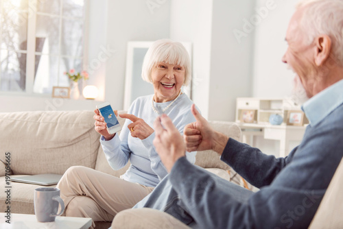 Great weather. Cheerful elderly couple sitting on the couch in the living room, checking the weather app and showing thumbs up, being happy about the weather