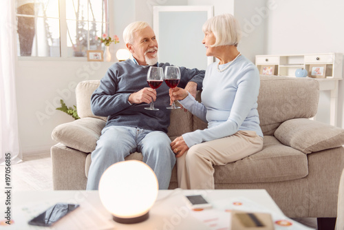 Romantic evening. Pleasant elderly couple sitting on the couch and drinking wine during the celebration of their wedding anniversary while looking at each other lovingly