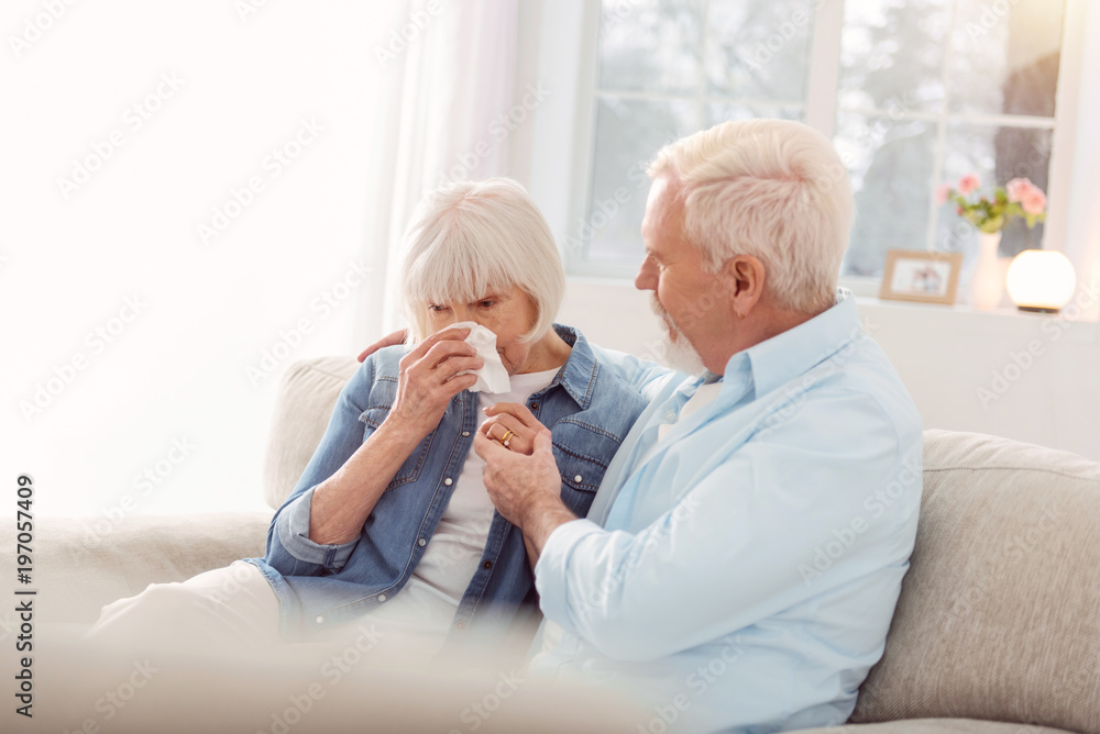 Do not cry. Caring elderly man sitting on the couch next to his wife and trying to cheer her up while she crying