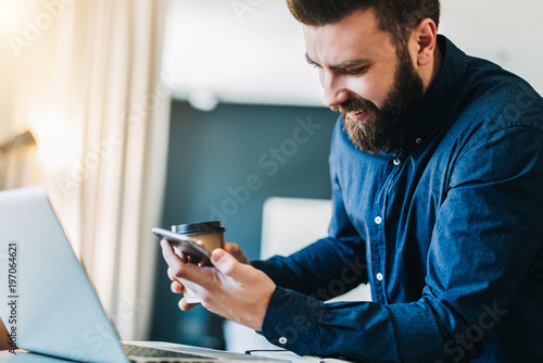 Smiling bearded businessman sitting at table in front of computer, using smartphone.Freelancer works home, distance work