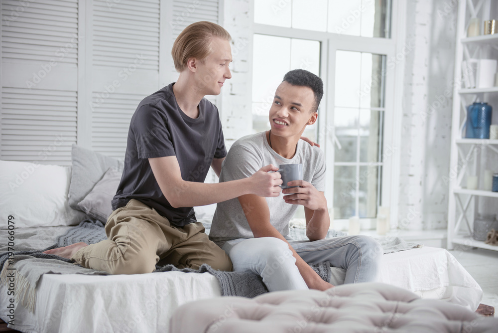 Everyday with you. Satisfied merry gay couple posing on bed while admiring each other and carrying cup Everyday with you. Satisfied merry gay couple posing on bed while admiring each other and carrying cup