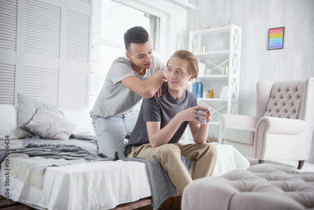 Sincere affection. Loving pleased gay couple posing in bedroom while talking and drinking coffee Sincere affection. Loving pleased gay couple posing in bedroom while talking and drinking coffee