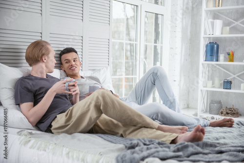 Life together. Positive energetic gay couple lying on bed while staring at each other and holding cups