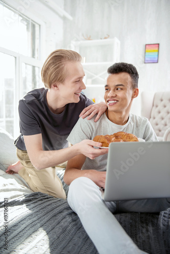 Best breakfast. Jovial merry gay couple sharing croissant while posing on bed and smiling