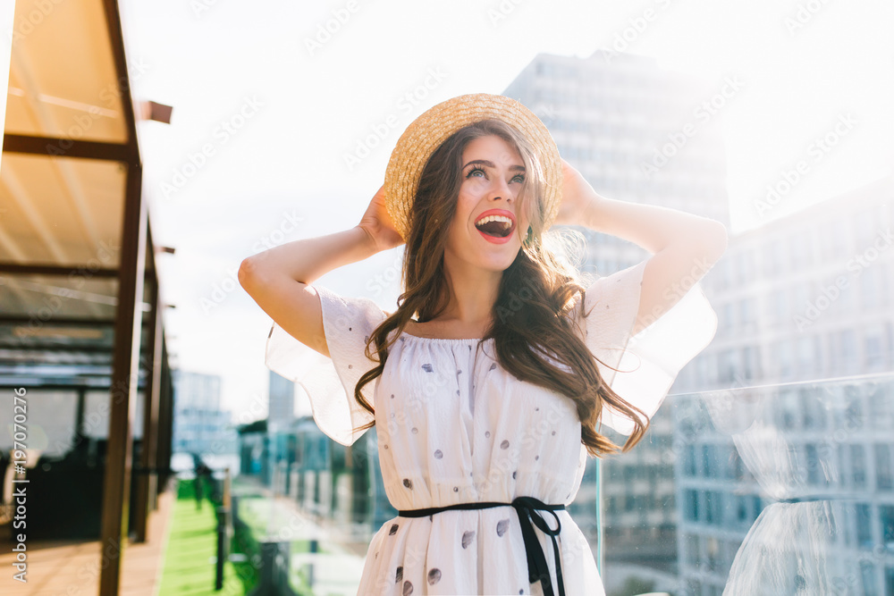 Fototapeta premium Happy girl with long hair in hat stands on the terrace on balcony . She wears a white dress with bare shoulders and red lipstick . She is smiling to the sky.