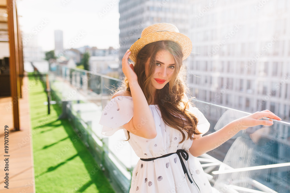 Pretty  girl with long hair in hat is enjoying  on the terrace on balcony . She wears a white dress with bare shoulders and red lipstick . She is looking to the camera.