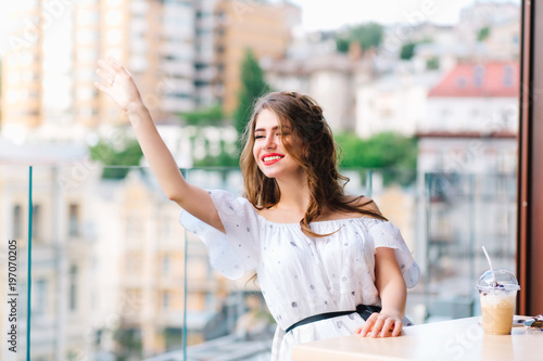 Free Download Beautiful girl with long hair is sittting at  table on the terrace in cafe. She wears a white dress with naked shoulders and red lipstick . She is calling for waiter. - GFXLoad