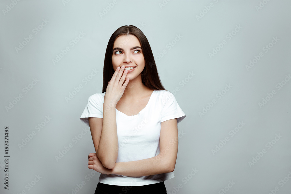 human emotions. Portrait of the young beautiful woman brunette with long hair. girl poses on a white background, smiles broadly, having covered a mouth with a hand. expresses confusion and shyness. human emotions. Portrait of the young beautiful woman brunette with long hair. girl poses on a white background, smiles broadly, having covered a mouth with a hand. expresses confusion and shyness.