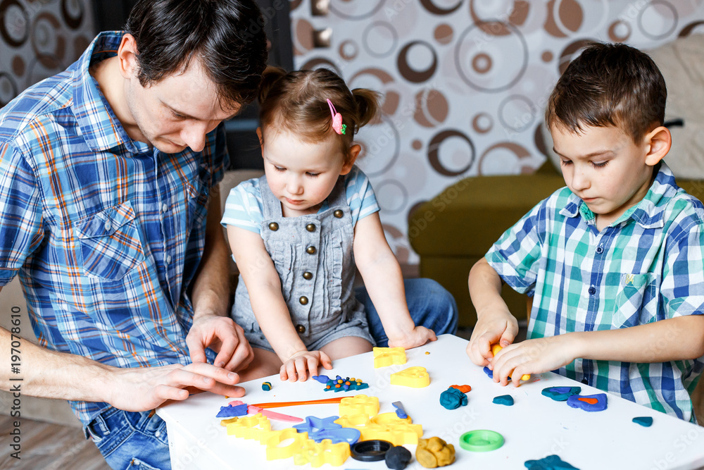 Fototapeta premium Picture of a father, daughter and son playing with color play dough and cutters. Having fun with colorful modeling clay. Creative kids molding at home. Children play with plasticine or dough.
