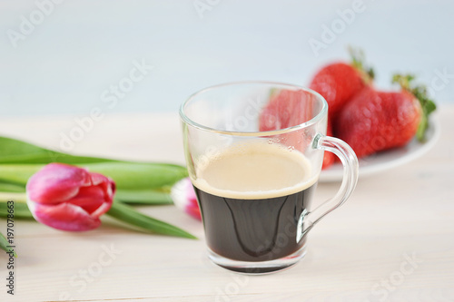 Composition with a cup of coffee and flowers. In a transparent cup of black coffee surrounded by pink tulips. In the background is a plate of strawberries. Light background. Close-up.