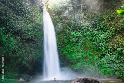 Huge fast waterfall hidden in mountain forest with person back view swimming. Deep jungle forest with bright green foliage, outdoor nature landscape, travel Bali, Indonesia lifestyle photography