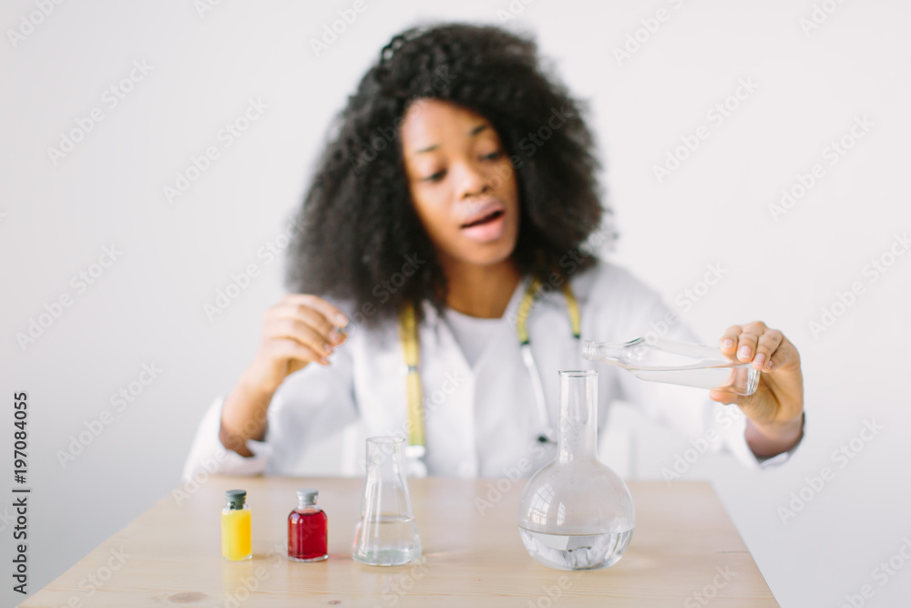 Young beautiful African American girl doctor in a white coat with a stethoscope. sitting at a table with reagent flasks on white background. Researcher researching in the laboratory Young beautiful African American girl doctor in a white coat with a stethoscope. sitting at a table with reagent flasks on white background. Researcher researching in the laboratory