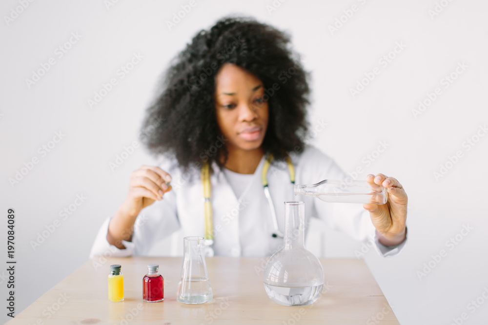 Young beautiful African American girl doctor in a white coat with a stethoscope. sitting at a table with reagent flasks on white background. Researcher researching in the laboratory Young beautiful African American girl doctor in a white coat with a stethoscope. sitting at a table with reagent flasks on white background. Researcher researching in the laboratory