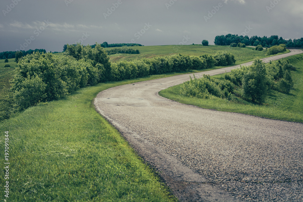 Dramatic green landscape with asphalt road in towards mountains under cloudy sky.