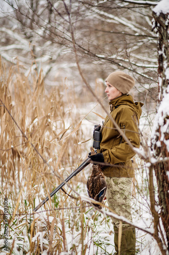 Female hunter in camouflage, armed with a rifle, standing in a snowy winter forest with duck prey