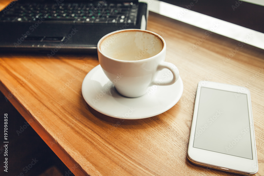 Drinking coffee while using a laptop in cafe. Finished coffee with phone and laptop in font of the window Drinking coffee while using a laptop in cafe. Finished coffee with phone and laptop in font of the window