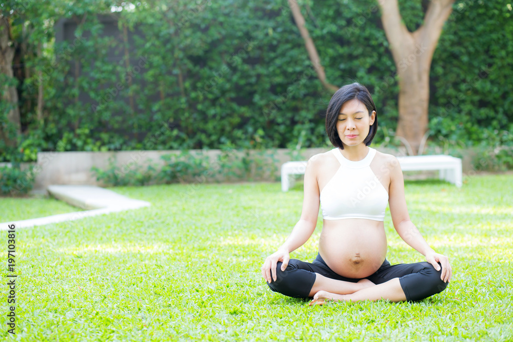 Beautiful pregnant asian young woman relax in the park, girl with belly sitting with crossed leg on grass and yoga exercise for wellness, copy space.