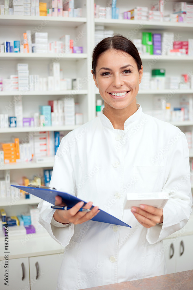 Medicine, pharmaceutics, health care and people concept - Laughing young pharmacist holding a clipboard and box of medications. Looking at camera