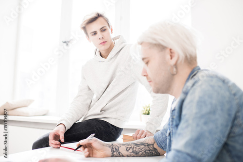 Young Caucasian male student sitting on desk and listening attentively to his adult college mate with tattooed hands