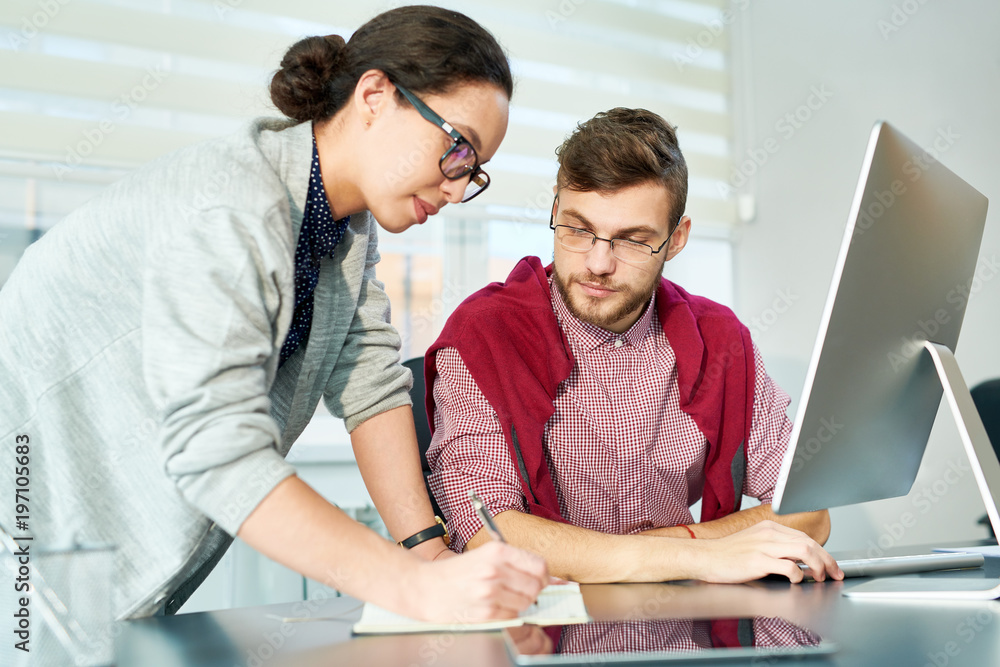 Attractive female office worker writing new data in notepad, young bearded colleague checking her while sitting at computer Attractive female office worker writing new data in notepad, young bearded colleague checking her while sitting at computer