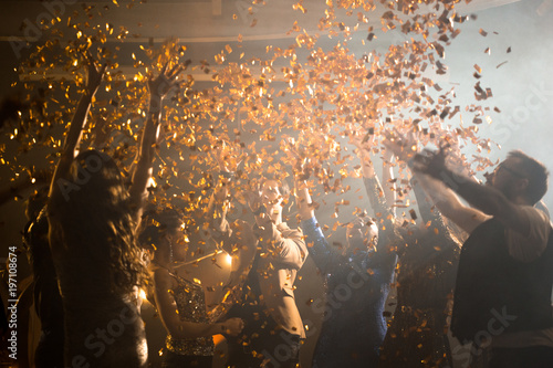 Joyful group of friends dancing to trendy music while hanging out at night club, golden confetti flying in air