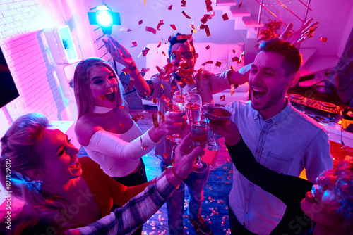 High angle view at group of happy friends enjoying holiday celebration at private house party raising glasses  and toasting under bursts of confetti