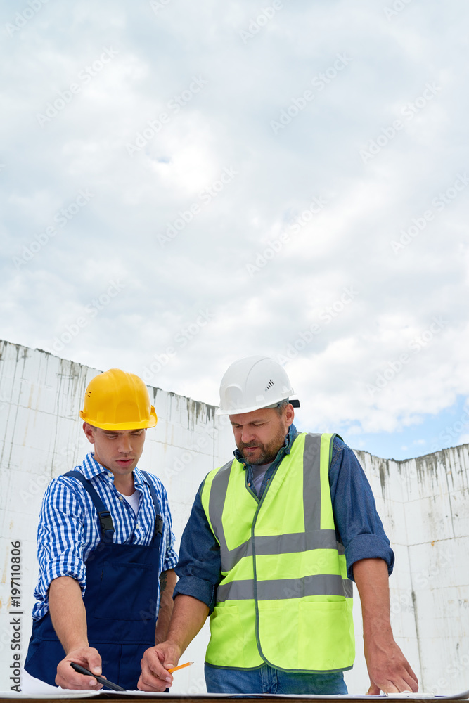 Waist up portrait of two construction workers discussing floor plans and engineering documentation on site making marks on blueprints, copy space Waist up portrait of two construction workers discussing floor plans and engineering documentation on site making marks on blueprints, copy space