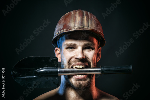 Construction worker concept - professional builder, repairman, engineer, miner in protective helmet holds shovel in teeth. Muscle sexy worker in harthat with shovel. Industrial worker with strict face photo