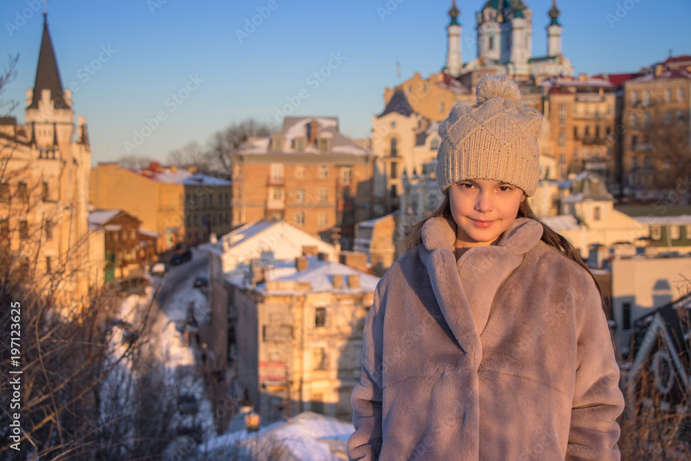 beautiful girl stands in a gray fur coat and a knitted beige hat on the background of houses