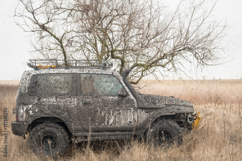  car in mud equipped for off-road driving stands in a dry grass under a tree