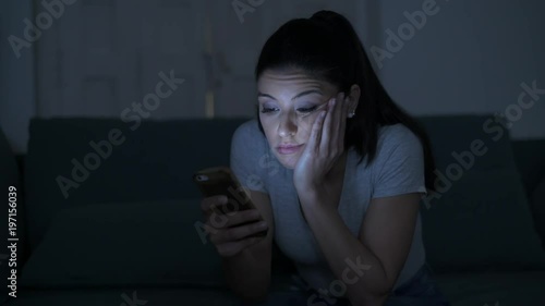 young attractive and tired hispanic 30s woman with mobile phone looking serious and bored using dating app sitting at apartment living room couch late night