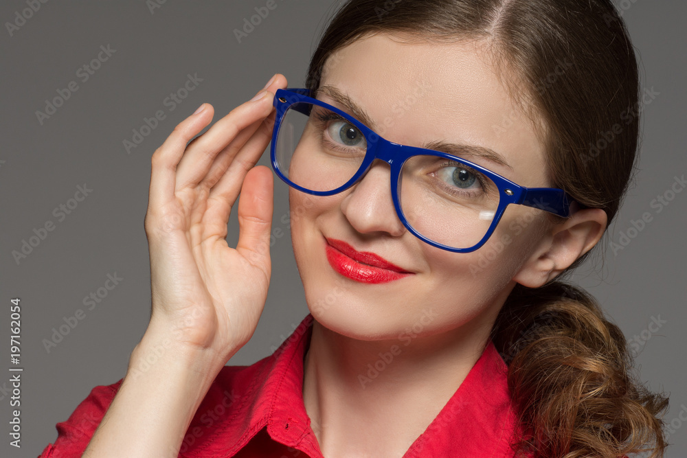 Beautiful brunette girl in a red shirt and wearing glasses smiling on a gray background