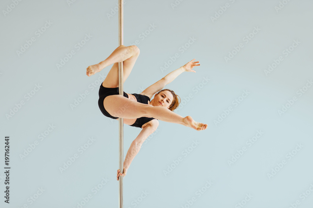 Fototapeta premium Beautiful pole dancer performing in the studio. Seria photo of middle aged red hair woman in black bra and shorts workout pole dance on air with different poses. Gray background.