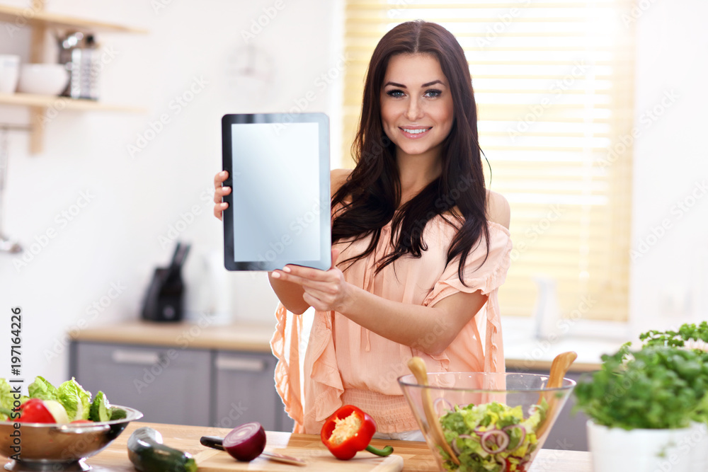 Happy woman preparing salad in modern kitchen and holding tablet