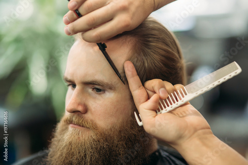 Bearded man getting a haircut by a professional hairdresser using comb and grooming scissors. Closeup view with shallow depth of field