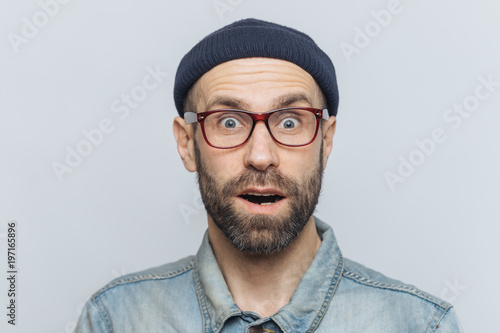 Foto Horizontal shot of amazed excited shocked man stares at camera with unexpected expression, keeps mouth opened, hears awful news, isolated over grey background